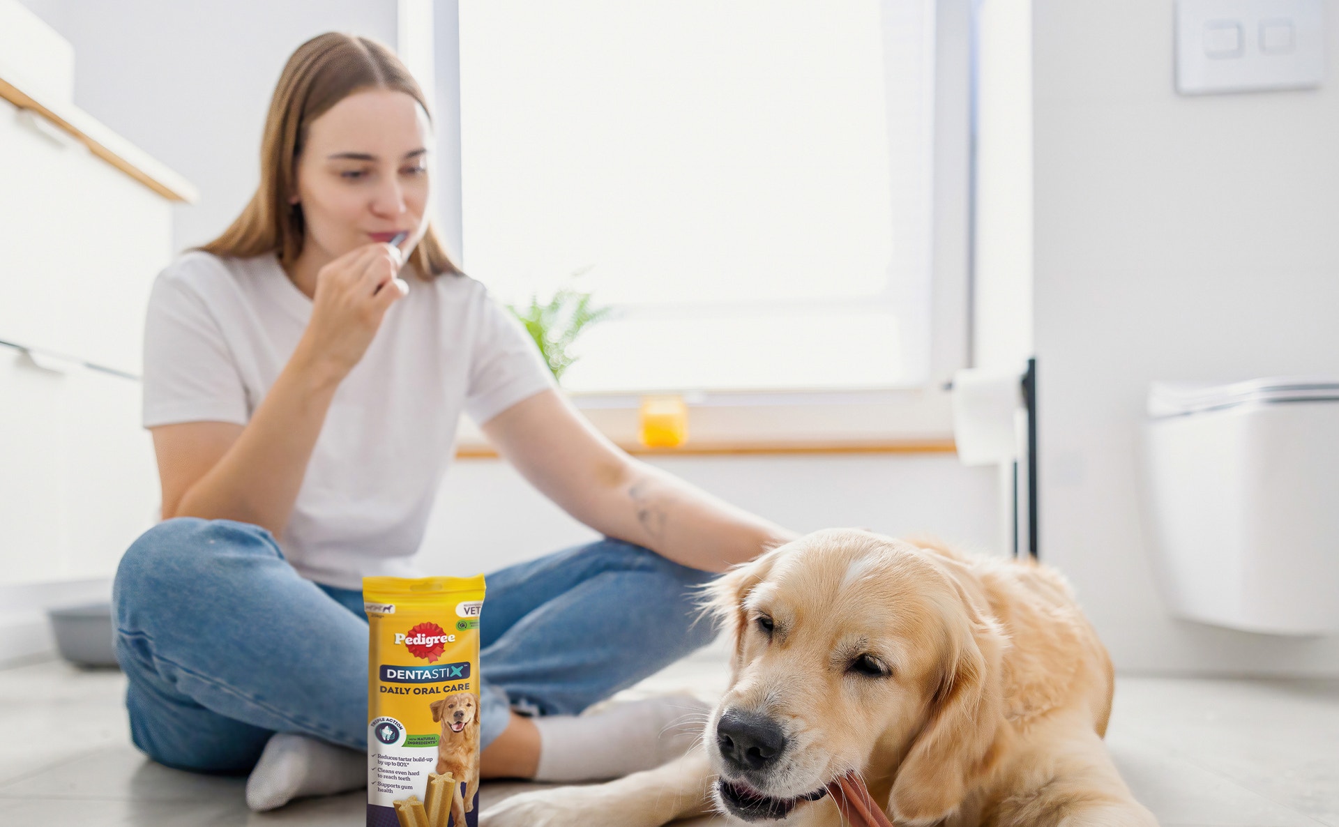 Eine junge Frau sitzt auf dem Boden mit einem Golden Retriever, der auf einem Pedigree Dentastix Kaustreifen kaut. Daneben liegt eine Packung Dentastix.